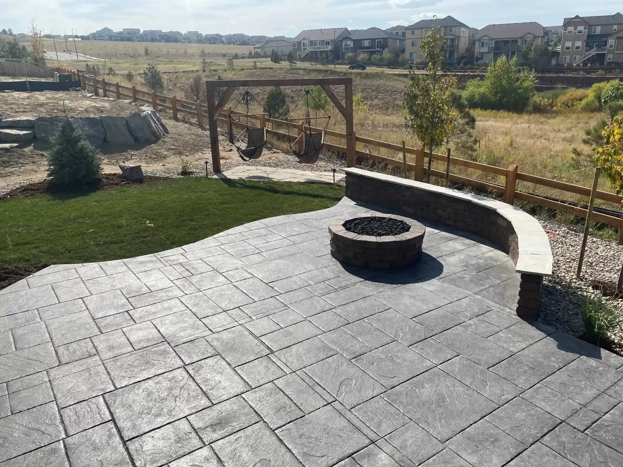 Stamped concrete patio with a fire-pit surround, curved seating wall, lawn, boulders, and an exposed sloped backdrop