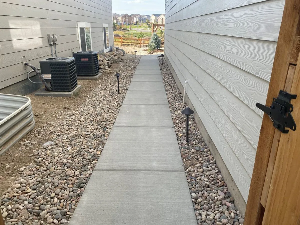 Narrow straight concrete walkway between two homes with decorative rock, utility equipment, and landscape lights