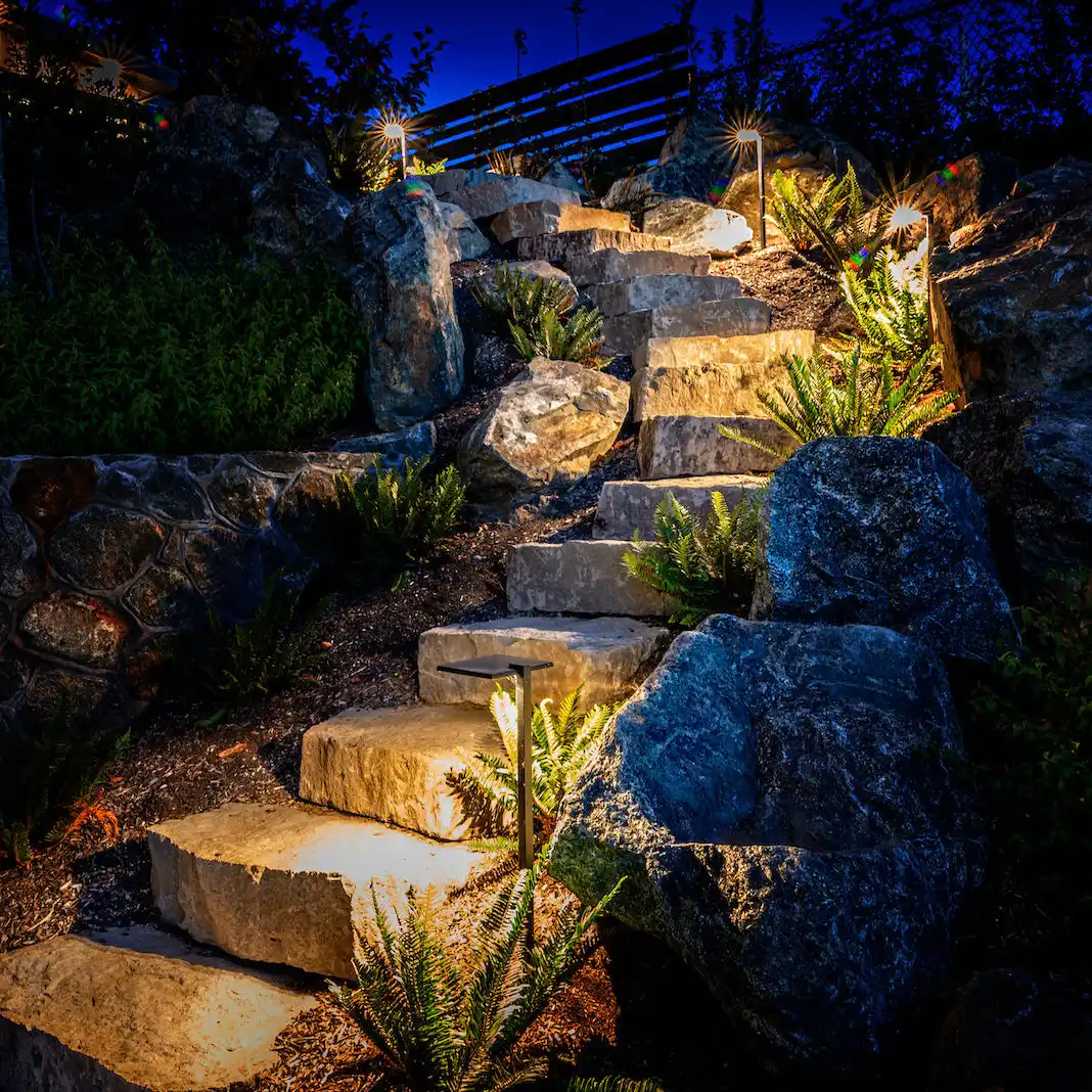 Stone steps through boulders and planting illuminated by visible landscape-lighting fixtures at night