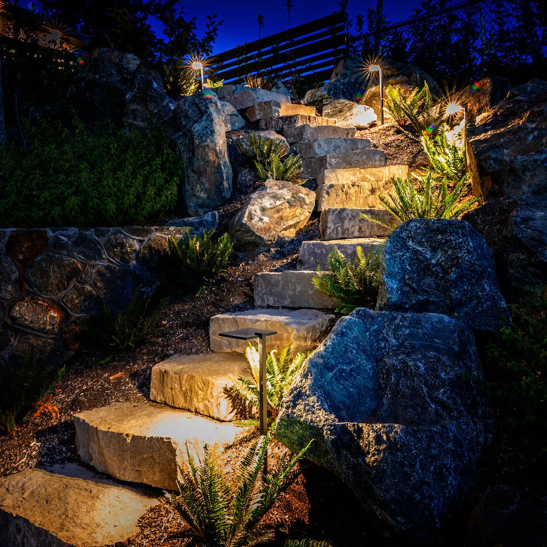 Stone steps through boulders and planting illuminated by visible landscape-lighting fixtures at night