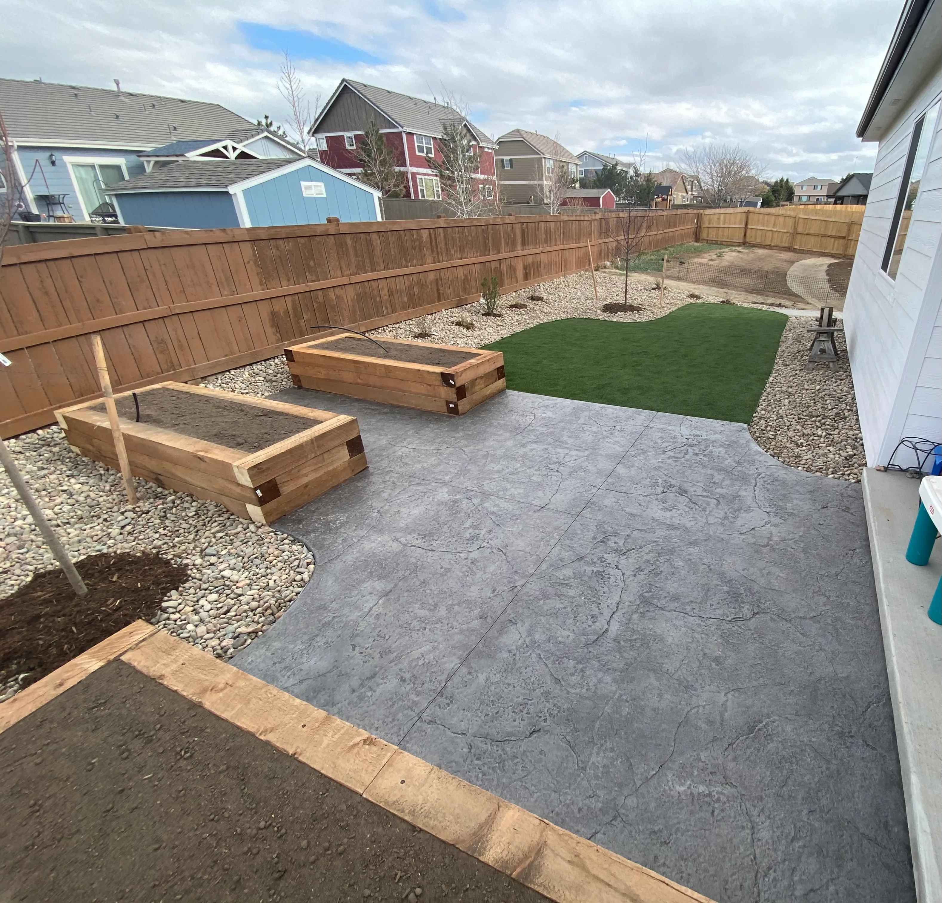 Stamped concrete patio connected to artificial turf, raised wood planters, decorative rock, and young trees