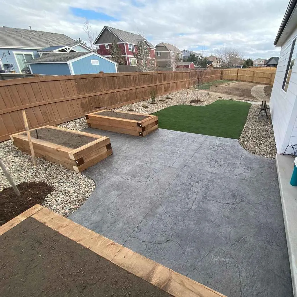 Stamped concrete patio beside raised wood planters, artificial turf, decorative rock, and young trees
