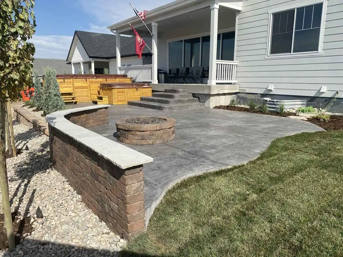 Newly completed backyard with a stamped concrete patio, a built-in fire pit, a block seating wall, fresh sod, young trees, and rock beds