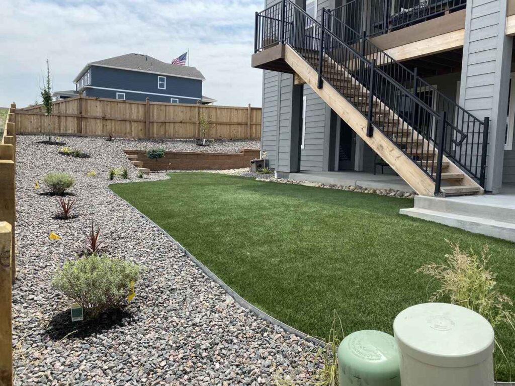 A landscaped backyard featuring a lush artificial turf lawn, decorative rock beds, and a wooden staircase leading to a deck in Parker, CO.