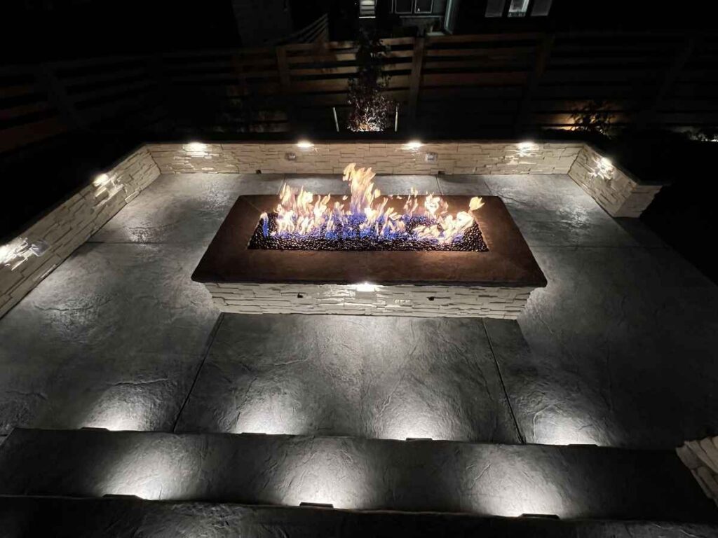 A modern fire pit surrounded by stylish stonework and illuminated by landscape lighting at night in Parker, CO.